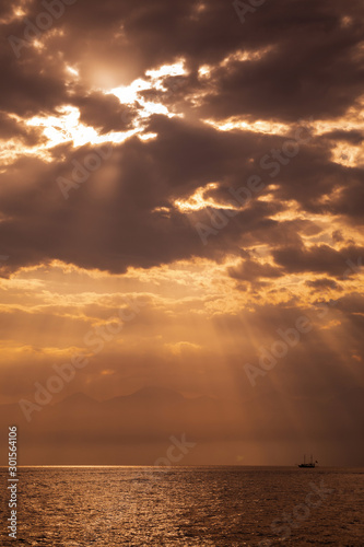 Amazing dramatic marine golden landscape. Sunny sea water, horizon line, cloudy sky with long sun rays transparenting through huge clouds, black silhouette of vintage ship in distance. Vertcial photo