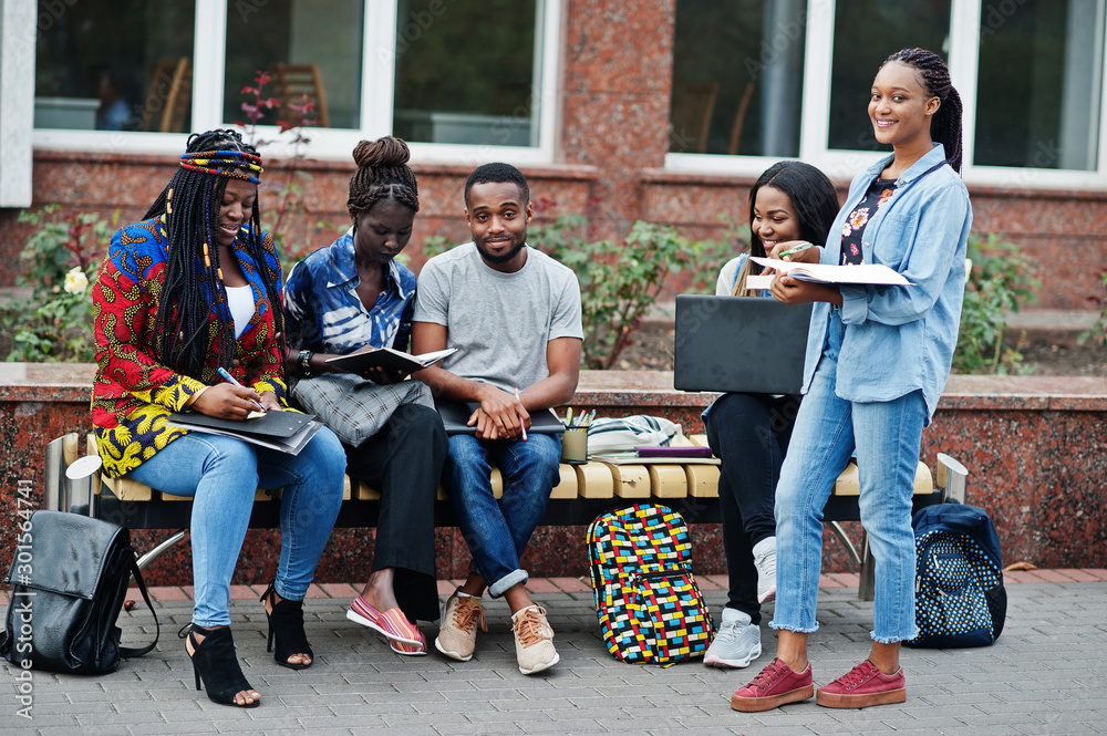 Fototapeta premium Group of five african college students spending time together on campus at university yard. Black afro friends studying at bench with school items, laptops notebooks.