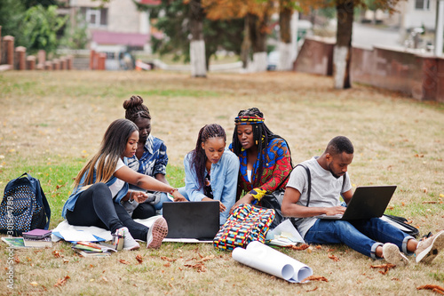 Group of five african college students spending time together on campus at university yard. Black afro friends sitting on grass and studying with laptops.