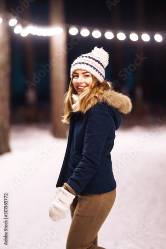 Portret of  beautiful girl is dressed in a blue jacket, in  knitted hat and mittens evening in the winter forest. Happy winter time in the forest. Festive garland lights.  Christmas, new year.