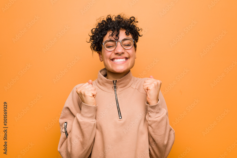 Young mixed african american woman against a brown background raising fist, feeling happy and successful. Victory concept.