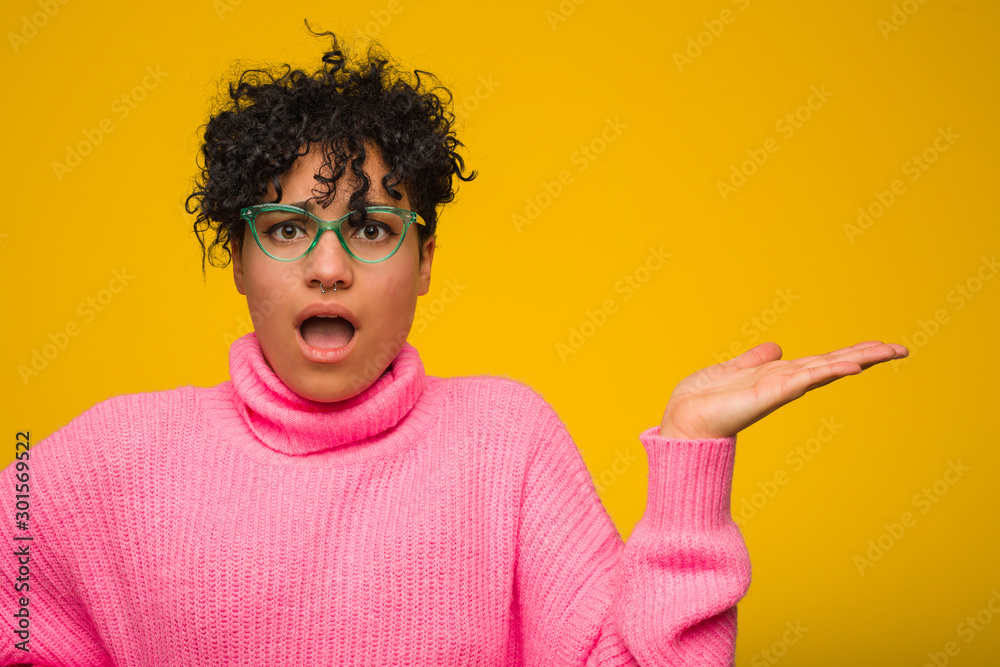 Young african american woman wearing a pink sweater impressed holding copy space on palm. Young african american woman wearing a pink sweater impressed holding copy space on palm.