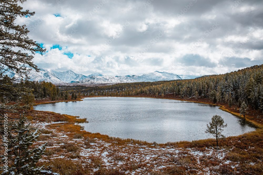 Panorama of mountain lake on background of beautiful snowy mountain peaks in cloudy weather. Mountain lake in the forest. View of lake on the background of blue sky