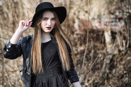 Young beautiful girl in a hat and with a dark make-up outside. Girl in the Gothic style on the street. A girl walks down the city street in a leather waistcoat with a phone. © alexkich