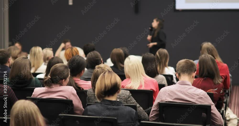 Educational researching training of modern technologies. Student science conference at university campus, woman speaker talking microphone for audience, view from auditorium sitting back to camera.