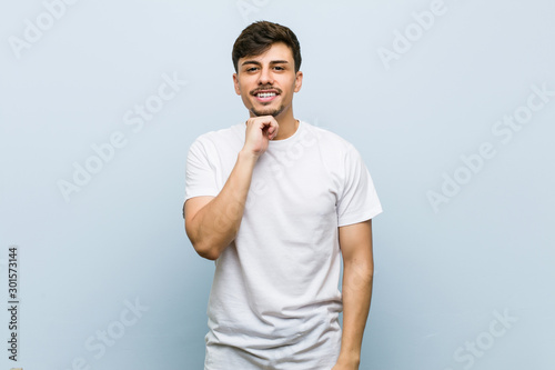 Young caucasian man wearing a white tshirt smiling happy and confident, touching chin with hand.