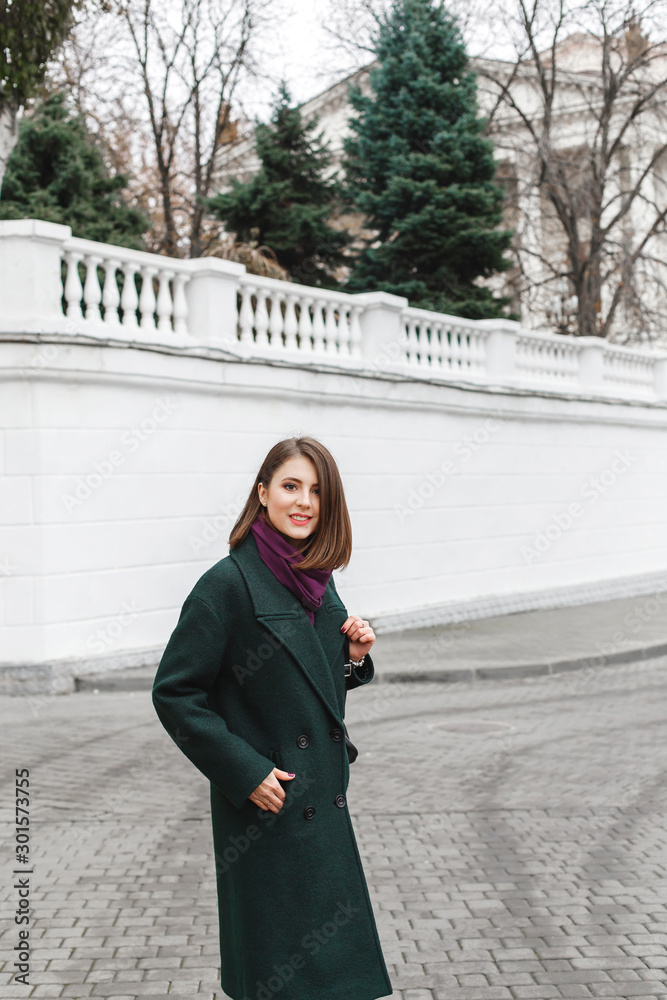 Beautiful young stylish brunette woman with short haircut, wearing green coat, purple scarf walking through the city streets. Trendy casual outfit. Street fashion. Everyday autumn, winter, spring look