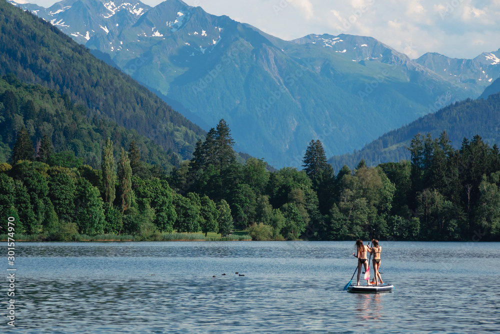 Two young beautiful girl-surfer riding on the stand-up SUP board in the clear waters of the Alpine mountains on the background. Lake 