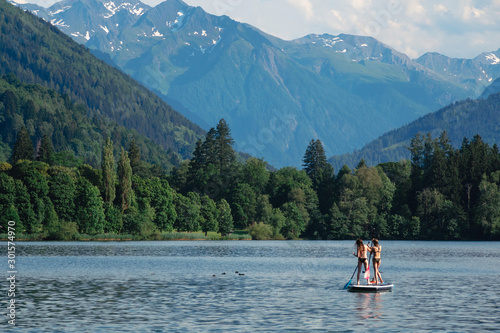 Two young beautiful girl-surfer riding on the stand-up SUP board in the clear waters of the Alpine mountains on the background. Lake "Zeller See" Austria