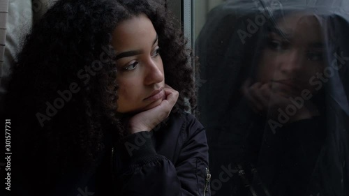 Sad depressed beautiful female mixed race African American girl teenager young woman teen wearing a black jacket sitting looking out of a window 