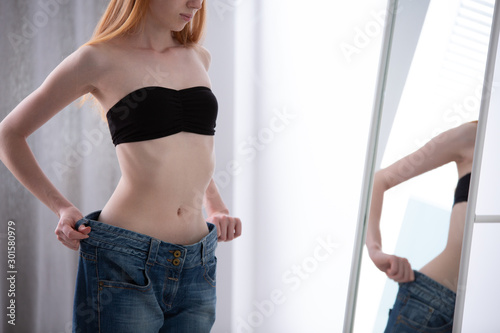Young anorectic girl wearing too big trousers and checking her reflection in the mirror