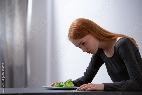 Redhead teenage girl on diet sitting at table and looking at plate with lettuce photo