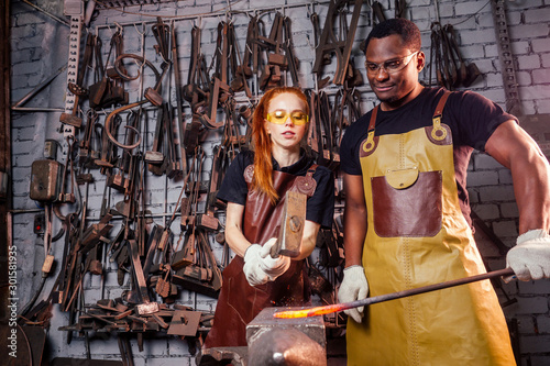 couple in love redhaired ginger young european woman and afro-american man wearing leather apron working blacksmith workshop.small family international business concept