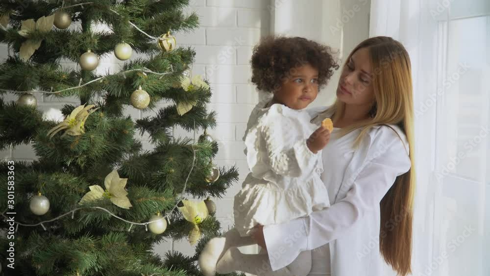 Happy mother with little daughter dressed in white sitting by the Christmas tree and eat cookies Happy mother with little daughter dressed in white sitting by the Christmas tree and eat cookies