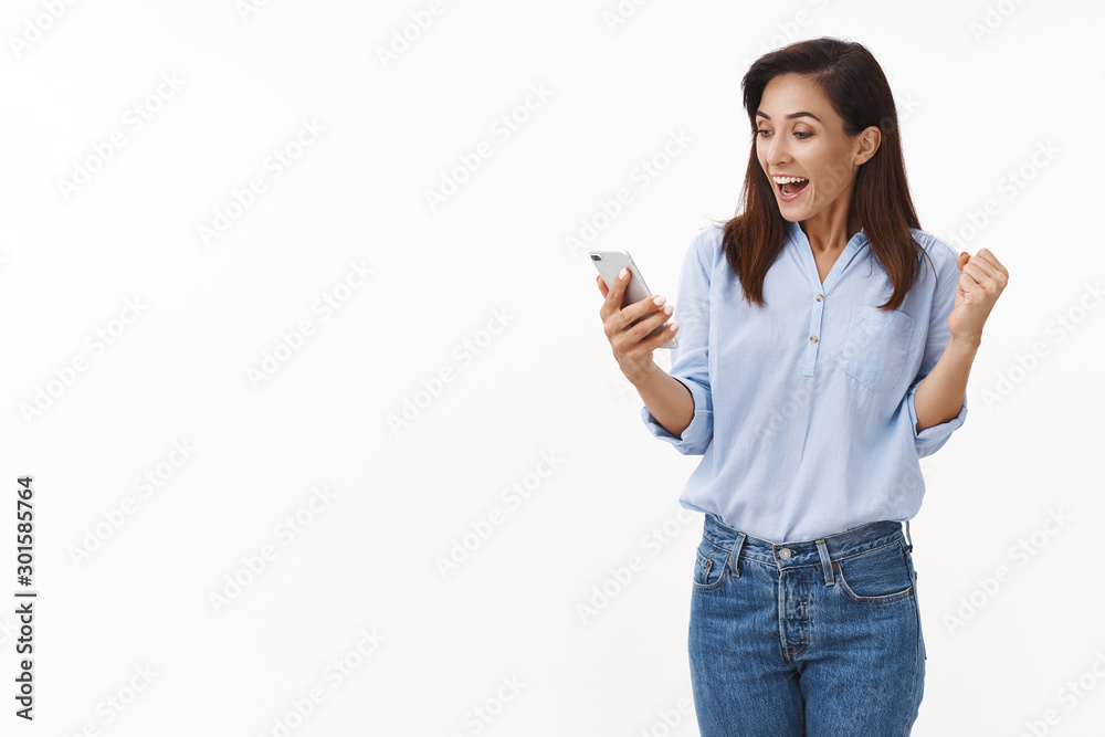 Cheerful triumphing adult 30s woman fist pump while reading excellent news, smiling broadly hold smartphone, look mobile phone display rejoicing, win online lottery, stand white background Cheerful triumphing adult 30s woman fist pump while reading excellent news, smiling broadly hold smartphone, look mobile phone display rejoicing, win online lottery, stand white background