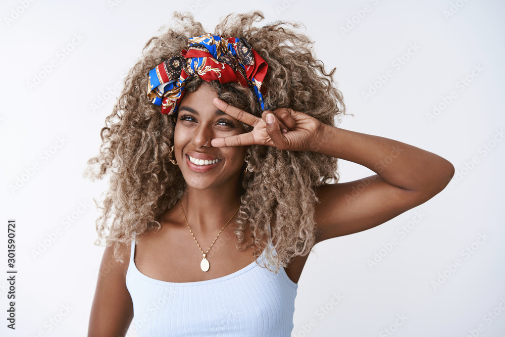 Naklejka premium Beauty, wellbeing and fashion concept. Cheerful, carefree african-american blond curly female in stylish headband, piercing and top, show peace, victory sign over eye send happy and joyful vibes