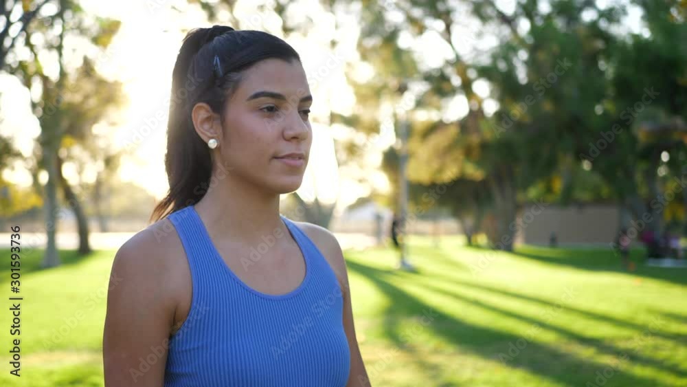 A pretty young hispanic woman smiling and working out in the park with dumbbells doing shoulder raises to increase her deltoid muscle strength. A pretty young hispanic woman smiling and working out in the park with dumbbells doing shoulder raises to increase her deltoid muscle strength.