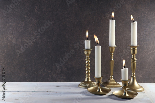 Burning candles in vintage metal candlesticks on white wooden table against dark stone background with copy space..