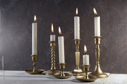 Burning candles in vintage metal candlesticks on white wooden table against dark stone background..