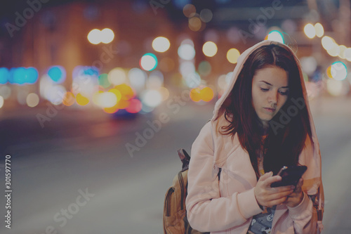 A beautiful girl in a pink sweater with a hood stands against the backdrop of the night city with a smartphone in her hand. Soft focus, colored highlights in the background.