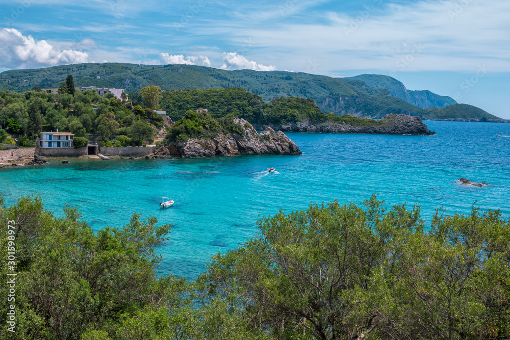 Landscape with turquoise calm sea water, mountain with rocky hillside covered with green trees and bushes and caves, cruise touristic boats and clouds on the sky. Corfu Island, Greece. 