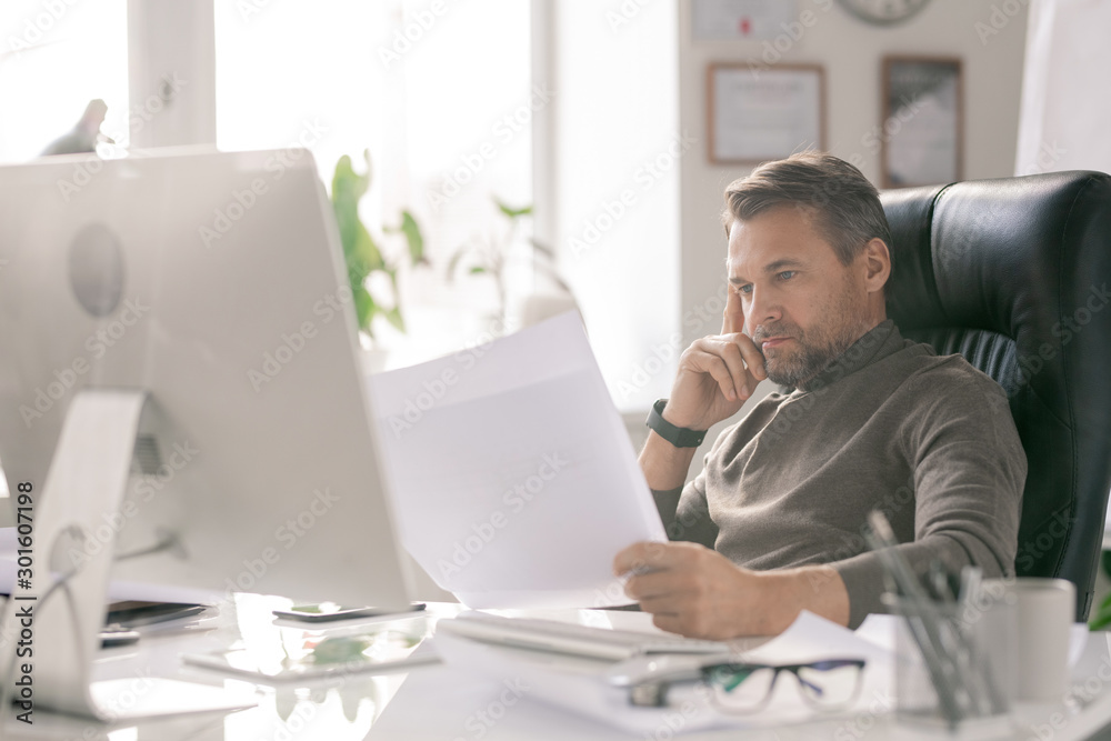 Mature serious architect looking at sketches while checking them by workplace