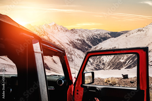 Winter red car on the road. Landscape of mountain peaks full of snow and sunset. Beautiful orange sky and Christmas time. Copy space.Winter road.