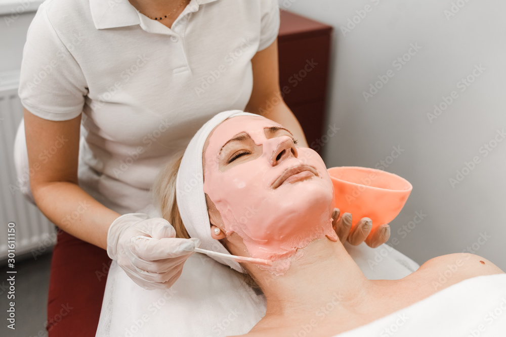 Obraz premium Doctor beautician applies anti-aging mask to woman face. Girls lies on a cosmetology procedure. Facial mask close-up