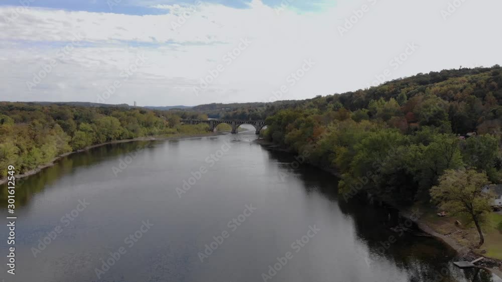 Colorful fall foliage on trees along a peaceful river. Homes on banks of river surrounded by autumn leaves. Aerial view from drone over water approaching arched bridge in distance. Colorful fall foliage on trees along a peaceful river. Homes on banks of river surrounded by autumn leaves. Aerial view from drone over water approaching arched bridge in distance.