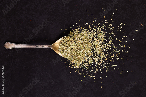 Crushed oregano scattered in an iron spoon on a black table. Concept, copy space.