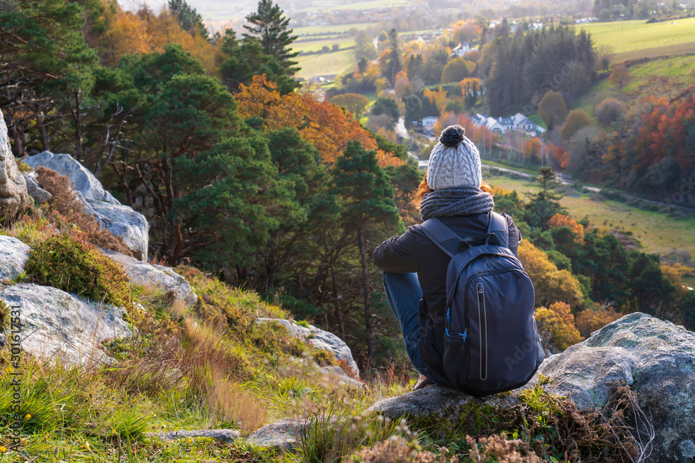 Fototapeta premium Young woman hiker wearing warm and cozy clothing, resting on a rock in Dublin Mountains, Ireland on a lovely fall day and enjoying the autumnal view from above. Irish autumn scenery.
