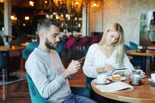 Serious man scrolling in smartphone on background of blonde girl making notes