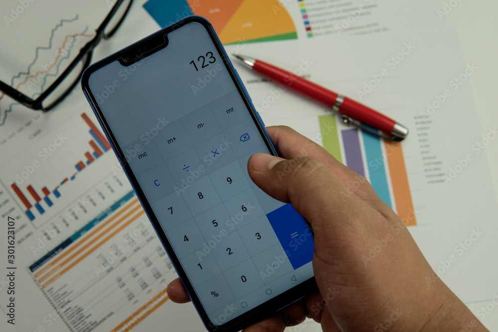 Fototapeta premium Adult male using phone as calculator with graph chart report document, pen and eye glasses on background. Finance banking concept.
