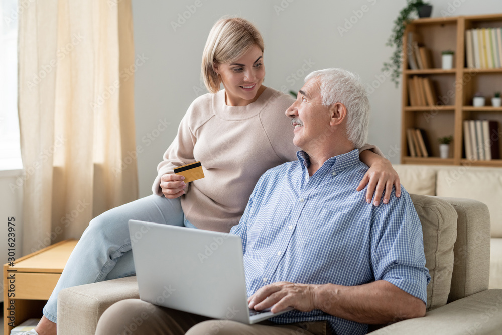 Pretty casual woman with credit card looking at her retired father with laptop Pretty casual woman with credit card looking at her retired father with laptop