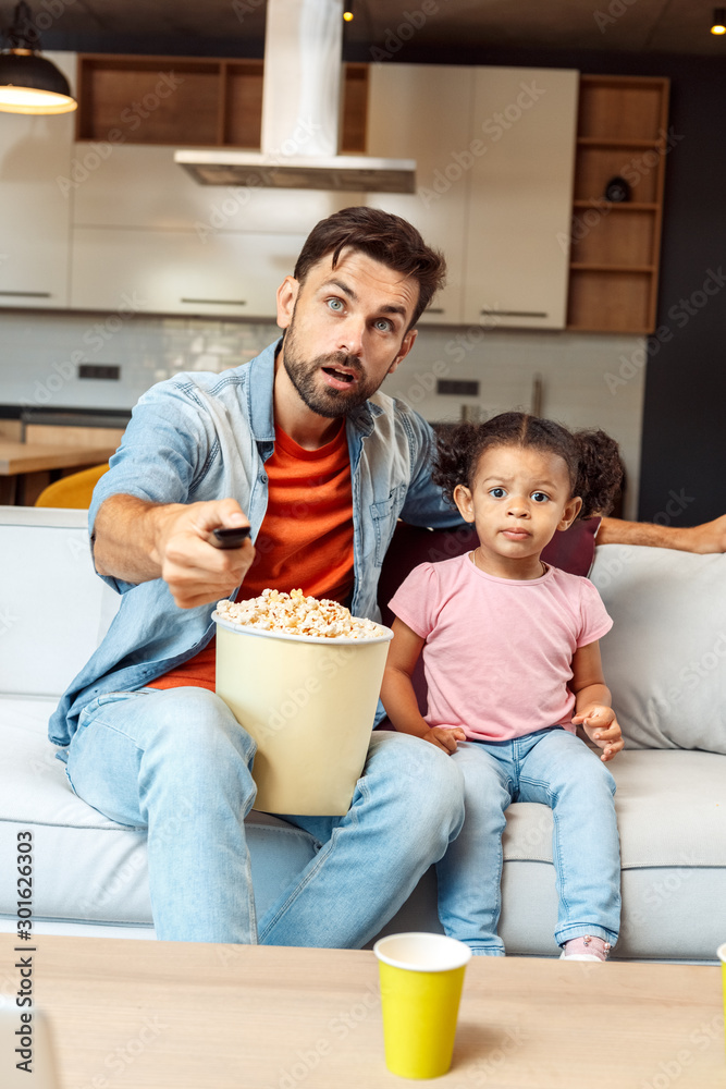 Mixed Race Family. Father and daughter sitting on sofa at home watching tv together eating snacks increasing volume looking at screen shocked Mixed Race Family. Father and daughter sitting on sofa at home watching tv together eating snacks increasing volume looking at screen shocked
