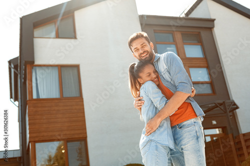 Mixed Race Marriage. Young man and woman standing outdoors near new house hugging cheerful in sunlight