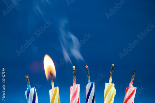 Colorful and fun birthday candles are being blown at a party. One candle is still lit and the other candles have already been blown out letting out an aromatic smoke. Blue background. Celebration
