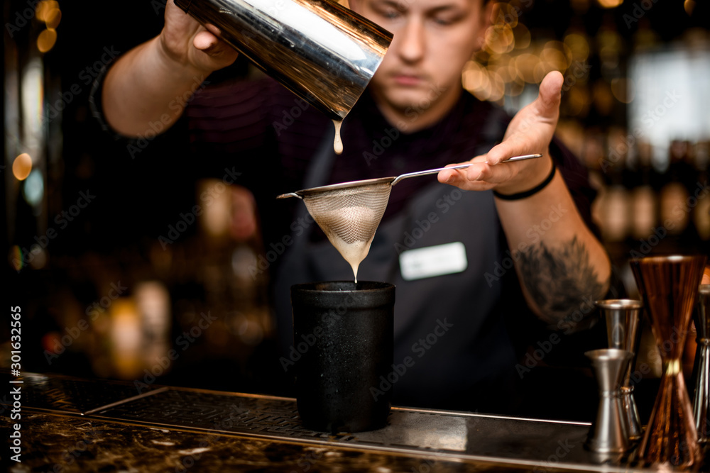 Bartender pouring a alcoholic drink from the steel shaker to the black glass through the sieve