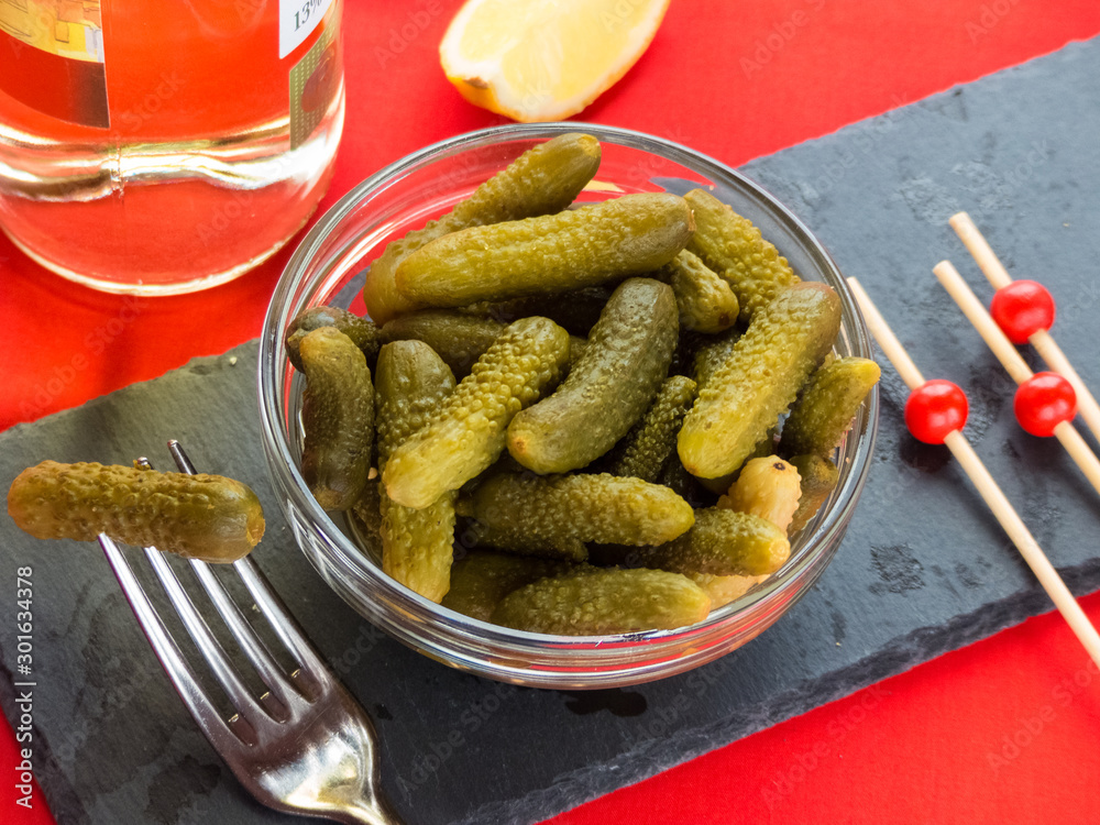 Gherkins in a crystal bowl on a slate table with toothpicks, metal fork, white wine and lemon in a red background. Gherkins in a crystal bowl on a slate table with toothpicks, metal fork, white wine and lemon in a red background.