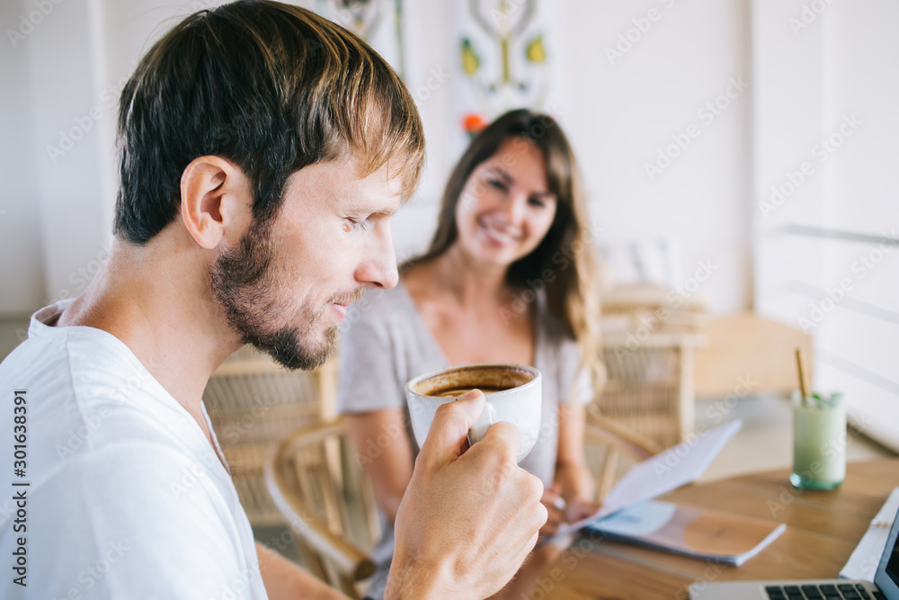 Side view of positive gladden hipster guy holding coffee cup in hands and enjoying aroma and taste of hot morning drink, Caucasian young man blowing on caffeine beverage during spare time indoors Side view of positive gladden hipster guy holding coffee cup in hands and enjoying aroma and taste of hot morning drink, Caucasian young man blowing on caffeine beverage during spare time indoors