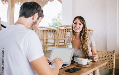 Portrait of cheerful hipster girl sitting at cafeteria table with milk fruit cocktail smiling at camera while male friend typing text for copywriter exchange on digital laptop with copy space