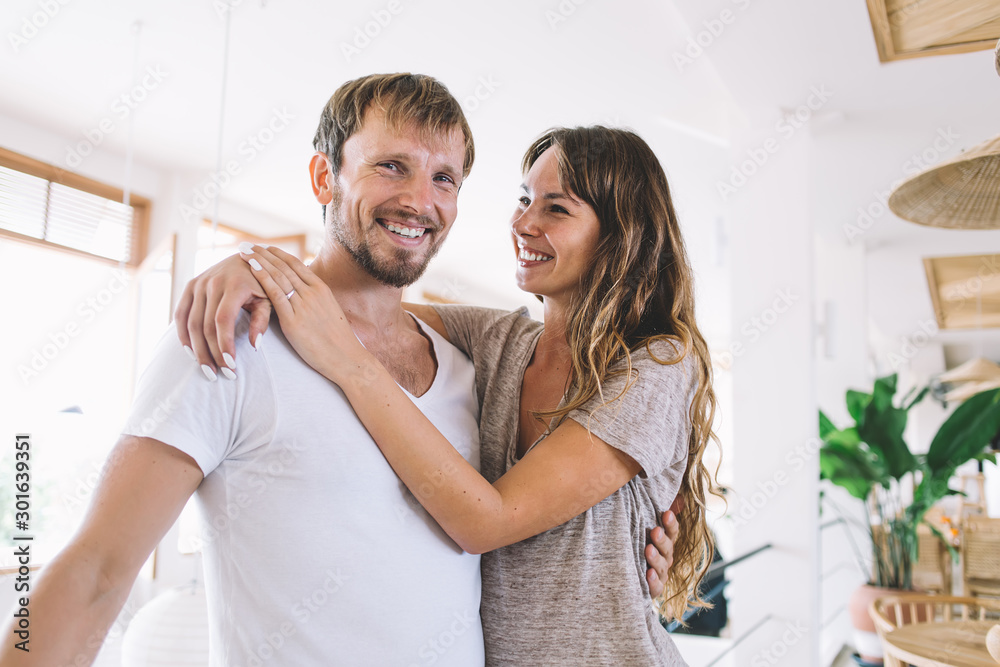 Half length portrait of cheerful bearded Caucasian hipster guy smiling at camera and enjoying joyful mood with lovely girlfriend hugging him, happy young marriage with amour feelings indoors Half length portrait of cheerful bearded Caucasian hipster guy smiling at camera and enjoying joyful mood with lovely girlfriend hugging him, happy young marriage with amour feelings indoors