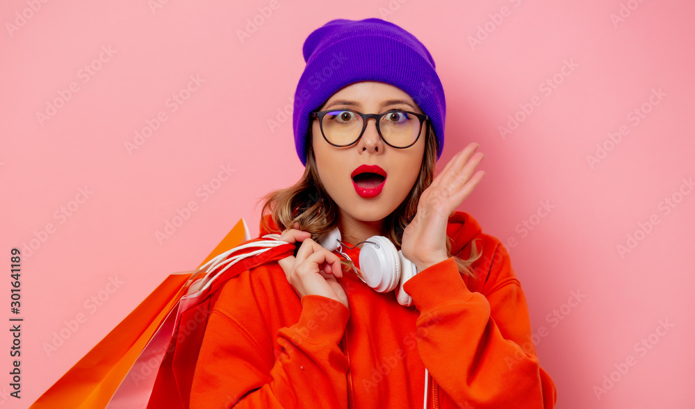Style girl in orange hoodie and purple hat with shopping bags on pink background Style girl in orange hoodie and purple hat with shopping bags on pink background