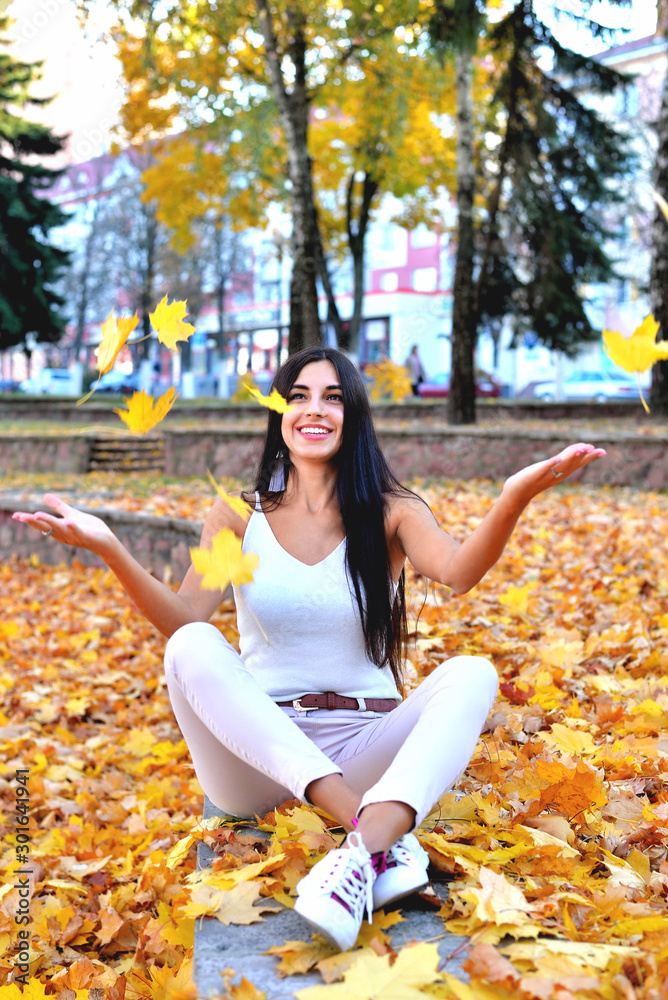 Fototapeta premium Beautiful girl in autumn park sitting on a concrete parapet catches falling maple leaves with her hands, a smile on her face, in white jeans and a blouse, against a background of trees 