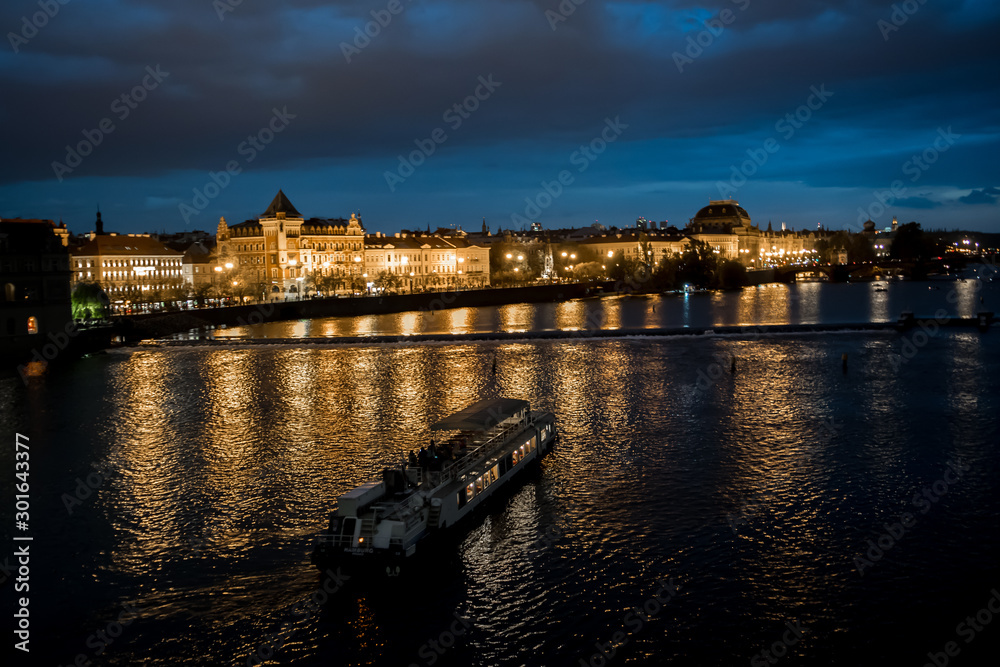 Illuminated Moldova River With Ship And Historic Buildings In The Night In Prague In The Czech Republic