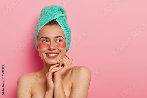 Smiling young woman wears cosmetic moisturizing patches under eyes, removes wrinkles, cares about complexion, wears soft towel on head, has cosmetics procedure, looks gladfully aside. Natural beauty