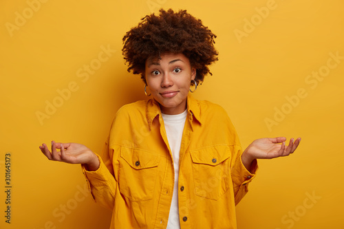 Puzzled clueless African American lady shrugs shoulders and expresses uncertainty, makes decision, wears fashionable clothing, spreads palms sideways isolated on yellow wall. Life perception, attitude photo
