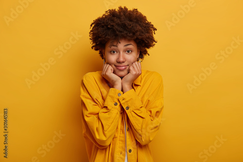 Satisfied curly haired woman with Afro hairstyle, keeps hands under chin, looks with sensual face expresion at camera, has naturral beauty, wears fashionable clothes, poses indoor over yellow wall