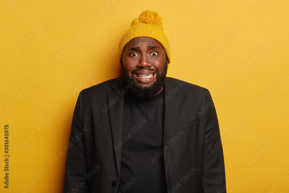 Nervous awkward black man with thick beard, feels bothered, admits his  mistake, clenches teeth, wears yellow knitted hat and formal suit, scared  of bad consequences, hears terrible news, isolated Stock Photo