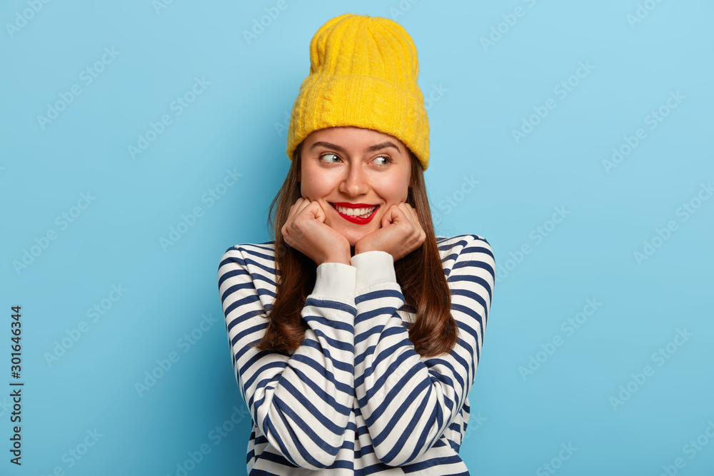 Beautiful brunette millennial woman keeps hands under chin, bites lips, has satisfied expression, wears yellow hat and striped jumper, dreams about romantic date, isolated over blue background Beautiful brunette millennial woman keeps hands under chin, bites lips, has satisfied expression, wears yellow hat and striped jumper, dreams about romantic date, isolated over blue background